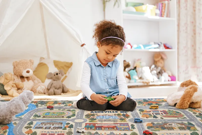 A girl sitting on an Asheville rug in a white playroom, holding a green toy car